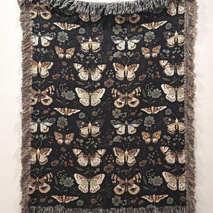 May include: A black woven throw blanket with a repeating pattern of brown and beige butterflies on a dark background. The blanket has a fringe border.