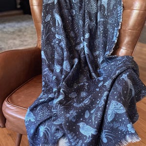 May include: A dark blue throw blanket draped over a brown leather armchair. The blanket features a repeating pattern of light blue forest creatures, plants, and insects. The blanket has a fringed edge.