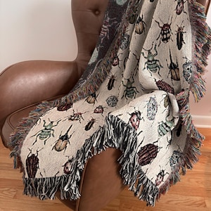 May include: A woven throw blanket featuring a repeating pattern of various colorful beetles and insects on a light cream background. The blanket has a textured fabric construction and is finished with decorative multi-colored fringe along the edges.