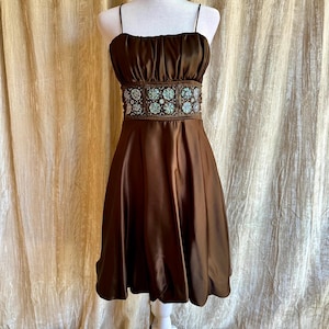 May include: A brown satin dress with spaghetti straps and a ruched bodice. The dress features a decorative belt with turquoise and silver embellishments. The skirt is a-line and falls to the knees. The dress is displayed on a white mannequin.