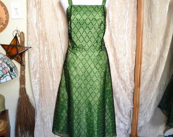 May include: A green knee-length dress with a diamond pattern overlay. The dress has a square neckline and spaghetti straps. The dress is displayed on a white mannequin. The background includes a mirror and a beige curtain.