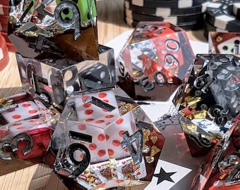 May include: Handmade resin dice with playing cards and gold leaf inclusions. The dice are various shapes and colours, including red, green, and clear, with black numbers. Poker chips are in the background.