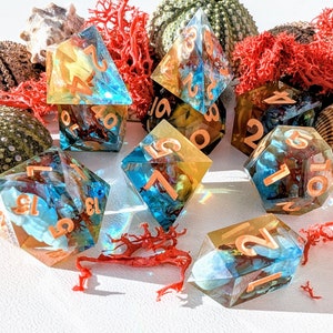 以下が含まれることがあります： 銅色の数字が書かれた、虹色の青と金のサイコロが8個。サイコロは、赤い珊瑚と貝殻が置かれた白い表面に散らばっている。