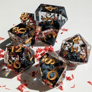 May include: A set of seven polyhedral dice for tabletop games. The dice are clear with black and red inclusions, and gold-coloured numbers. The dice are scattered on a white surface with red flower petals.