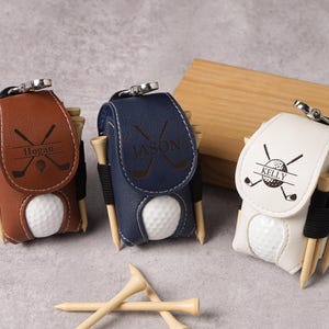 May include: Three personalized golf ball holders in brown, navy blue, and white leather. Each holder has a golf ball, tees, and a name. The brown holder says "Hogan", the navy blue holder says "Jason", and the white holder says "Kelly".