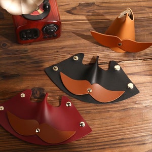 May include: Three leather mustache-shaped keychains in different colors: brown, black, and red. Each keychain has a brown leather mustache with gold accents.