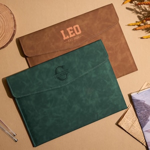 May include: Two leather document holders, one brown and one dark green, are displayed. The brown holder has the name "LEO" and the text "EST. 2020" printed on it. The green holder has a circular logo. The holders are on a tan surface.