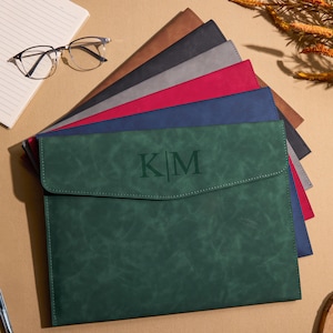 May include: A collection of leather document holders in various colors, including green, red, and blue. The green holder in the foreground features the initials "KM". A notebook, glasses, and a pen are also visible.