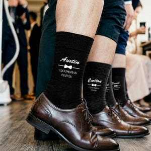 May include: Black crew socks featuring white printed text and a bow tie graphic. The text includes individual names such as Austin and Carlton, the title GROOMSMAN, and the date 08.09.26. The socks are worn with dark brown leather dress shoes.
