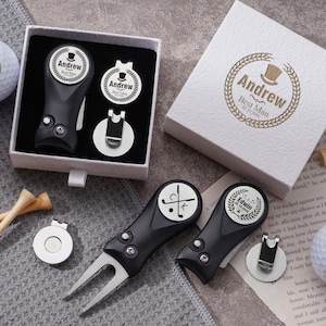 Könnte beinhalten: Ein Golf-Geschenkset mit weißen Golfbällen, Holztees und schwarzen Metall-Pitchgabeln mit silbernen magnetischen Ballmarkern. Die Artikel sind mit den Namen Andrew und Edwin, einem Zylinder-Symbol und Golfschläger-Grafiken personalisiert und in einer weißen, strukturierten Geschenkbox verpackt.