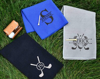 May include: Three golf towels in blue, gray, and black, with personalized embroidery. The blue towel has "STEVEN" and a golf tee. The gray towel has "PATRICK" and golf club graphics. A leather golf ball holder is also visible.