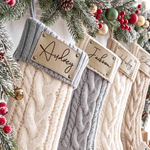 May include: A row of four knit Christmas stockings in cream and light gray, each featuring a cable knit pattern and a ribbed cuff. A rectangular tan leather tag is attached to each cuff with metal rivets, displaying the names Audrey, Jackson, Hailey, and Edward in black script.