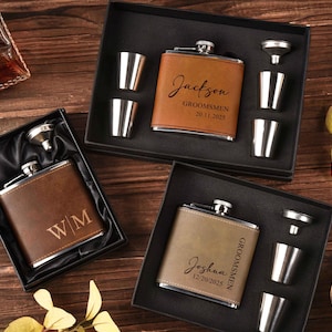 Könnte beinhalten: Drei personalisierte Lederflaschen in schwarzen Geschenkboxen, jeweils mit Schnapsgläsern und einem Trichter. Eine Flasche ist braun mit der Gravur "Jackson GROOMSMEN 20.11.2025". Eine weitere ist braun mit der Gravur "WM". Die dritte ist beige mit der Gravur "Joshua GROOMSMEN 12/20/2025".