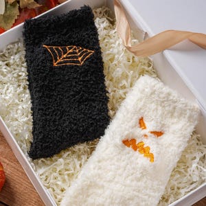 May include: A pair of fuzzy socks in a white gift box. One sock is black with an embroidered orange spider web, and the other sock is white with an embroidered orange jagged mouth design. The socks are nestled in white shredded paper filler.