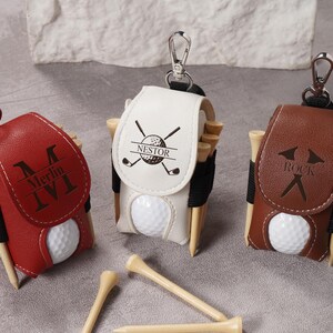 May include: Three golf ball holders in red, white, and brown leather, each with a golf ball and wooden tees. The red holder has "Merlin IVI" engraved, the white has "Nestor" and golf club graphics, and the brown has "Rock".