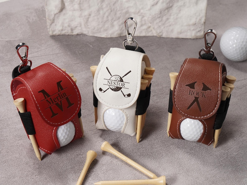 May include: Three golf ball holders in red, white, and brown leather, each with a golf ball and wooden tees. The red holder has "Merlin IVI" engraved, the white has "Nestor" and golf club graphics, and the brown has "Rock".