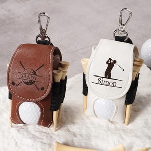 May include: Two personalized golf ball holders with attached keychains. One is brown with "Sandy" and golf club graphics, the other is white with a golfer silhouette and "Simon". Both have golf tees and a golf ball.