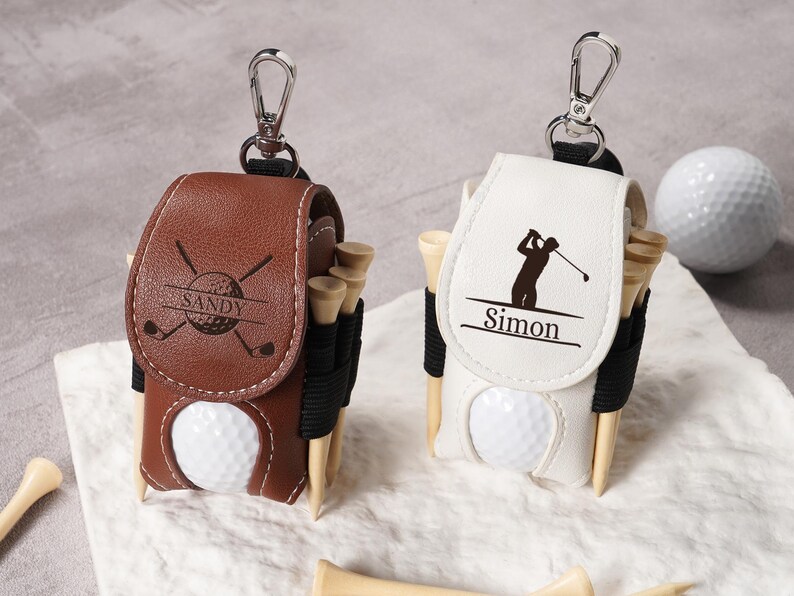 May include: Two personalized golf ball holders with attached keychains. One is brown with "Sandy" and golf club graphics, the other is white with a golfer silhouette and "Simon". Both have golf tees and a golf ball.