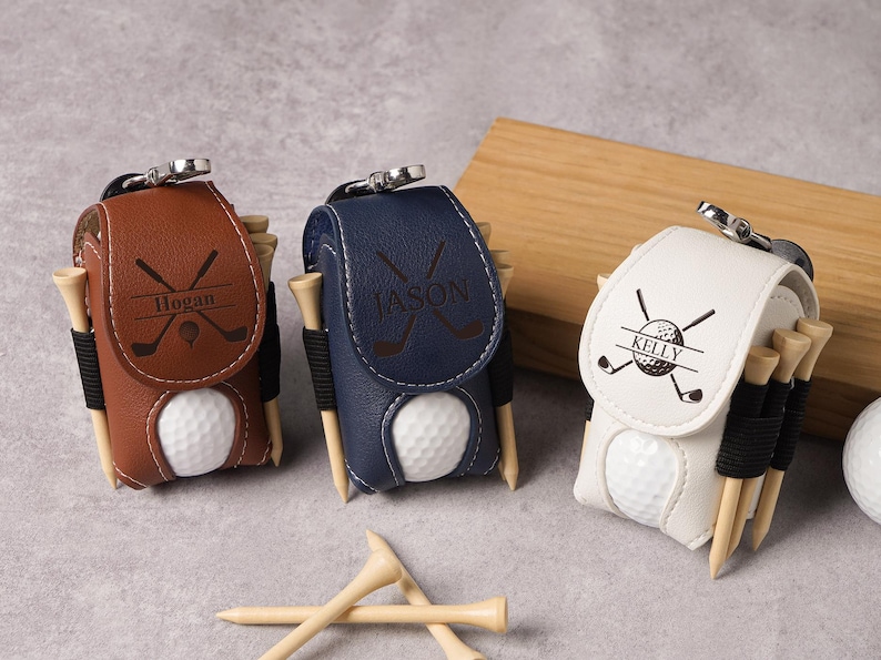 May include: Three personalized golf ball holders in brown, navy blue, and white leather. Each holder has a golf ball, tees, and a name. The brown holder says "Hogan", the navy blue holder says "Jason", and the white holder says "Kelly".