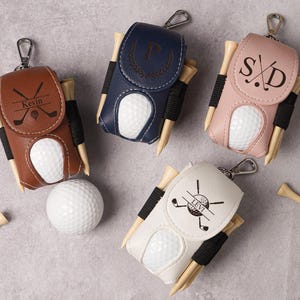 May include: Four personalized golf ball holders in brown, navy, pink, and white leather. Each holder has a golf ball, tees, and a silver clip. The holders are personalized with initials or names and golf-themed graphics.