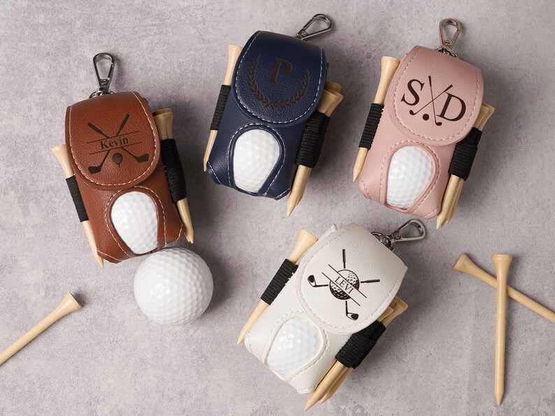 May include: Four personalized golf ball holders in brown, navy, pink, and white leather. Each holder has a golf ball, tees, and a silver clip. The holders are personalized with initials or names and golf-themed graphics.