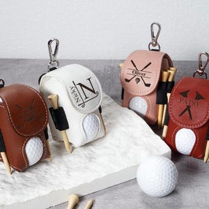 May include: Four personalized golf ball holders in brown, white, pink, and red leather. Each holder has a golf ball, tees, and a silver clip. The holders are personalized with names and initials.