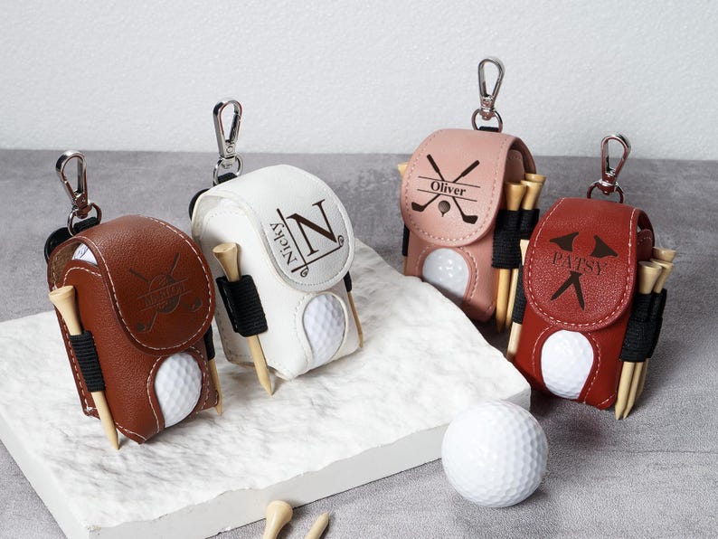 May include: Four personalized golf ball holders in brown, white, pink, and red leather. Each holder has a golf ball, tees, and a silver clip. The holders are personalized with names and initials.