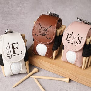 May include: Three personalized golf ball holders in white, brown, and pink leather. Each holder has a golf ball, wooden tees, and a silver clasp. The white holder has the name "Evan" and the letter "F". The brown holder has the name "Haley". The pink holder has the initials "E S".