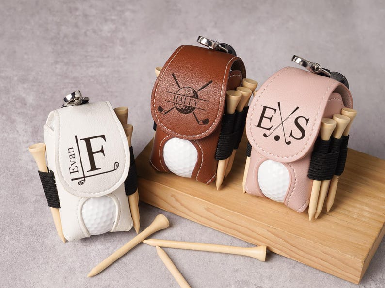 May include: Three personalized golf ball holders in white, brown, and pink leather. Each holder has a golf ball, wooden tees, and a silver clasp. The white holder has the name "Evan" and the letter "F". The brown holder has the name "Haley". The pink holder has the initials "E S".