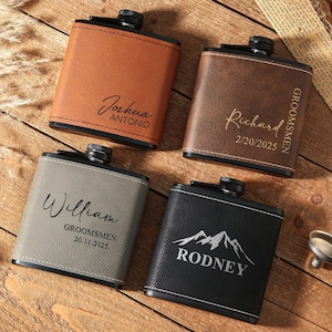 Peut inclure: Quatre flasques personnalisés en cuir marron, marron foncé, gris et noir. Chaque flasque porte un nom différent gravé, avec les mots "Groomsmen" et une date sur deux d'entre eux. Une flasque a un graphique de montagne et le nom "Rodney".