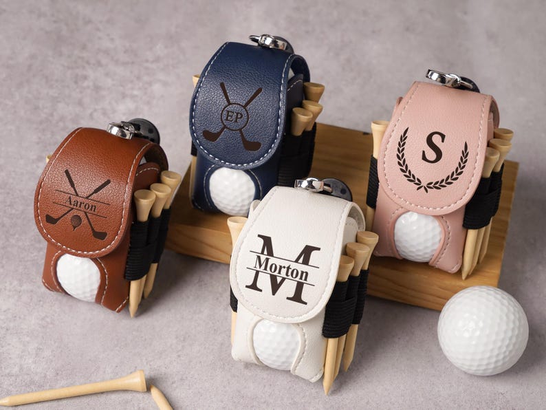 May include: Leather golf ball pouches in brown, navy, white, and pink, each with a golf ball and tees. The pouches have personalized initials or names, and crossed golf club designs. A golf ball and tees are also visible.