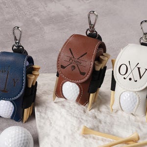 May include: Three personalized golf ball holders in navy blue, brown, and white leather. Each holder has a golf ball, tees, and a silver clip. The navy blue holder has the letters "K Y", the brown holder has the word "Primo", and the white holder has the letters "O X V".