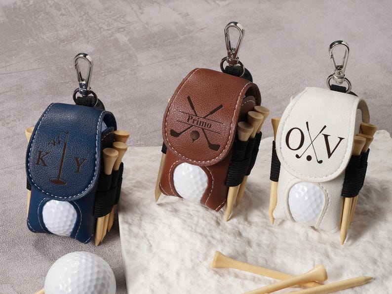 May include: Three personalized golf ball holders in navy blue, brown, and white leather. Each holder has a golf ball, tees, and a silver clip. The navy blue holder has the letters "K Y", the brown holder has the word "Primo", and the white holder has the letters "O X V".
