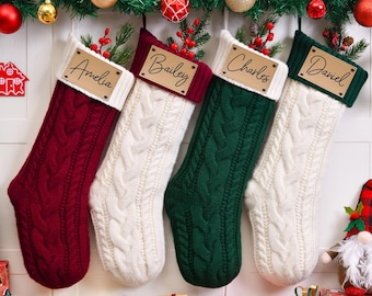 Op de afbeelding: Vier kabelgebreide kerstsokken in rood, wit en groen, elk met een gepersonaliseerd naamlabel. De sokken zijn versierd met een witte manchet en een bruin naamlabel van kunstleer. Feestelijke vakantiedecoratie.
