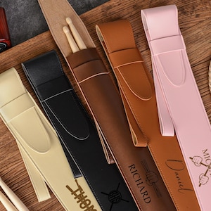 Puede incluir: Una variedad de estuches personalizados de cuero para baquetas en crema, negro, marrón y rosa. Cada estuche presenta un nombre y un gráfico de baquetas. Los estuches se exhiben sobre una superficie de madera, con baquetas visibles.