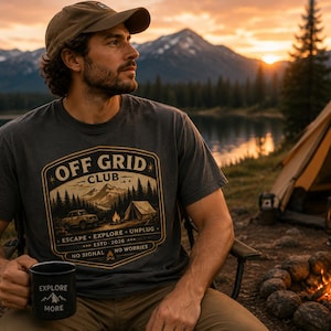 Op de afbeelding: Een antracietgrijs T-shirt met korte mouwen en een grafische print met de tekst OFF GRID CLUB, ESCAPE EXPLORE UNPLUG, ESTD 2026, NO SIGNAL NO WORRIES, en een bergkampeerscène. In één hand wordt een zwarte mok met de tekst EXPLORE MORE vastgehouden.