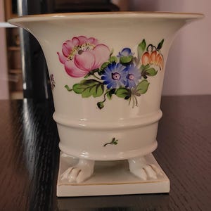 Peut inclure: Un vase en porcelaine blanche avec un bord évasé et une base carrée. Le vase est décoré d'un motif floral rose, bleu et orange. La base a deux pieds en forme de griffes. Le vase mesure environ 18 cm de haut.