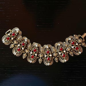 May include: Gold-toned bracelet with ornate filigree design. The bracelet features oval-shaped red gemstones and small white accents. The clasp is visible on the left side. The bracelet is displayed on a dark surface.