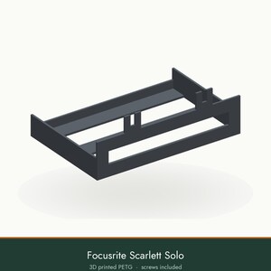 May include: A dark gray 3D printed PETG mounting bracket designed for a Focusrite Scarlett Solo audio interface. The rectangular frame features integrated support tabs and open sides, providing a structured housing for the device.