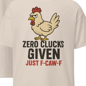 May include: Beige t-shirt featuring a cartoon chicken and the phrase "ZERO CLUCKS GIVEN JUST F-CAW-F". The chicken is light brown with a red comb and beak. The shirt has a crew neck and short sleeves.