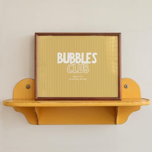 Könnte beinhalten: Gerahmter Druck mit den Worten "BUBBLES CLUB" in weißen Buchstaben auf gelbem Hintergrund mit vertikalen Streifen. Der Rahmen ist braun, und der Druck liegt auf einem gelben Regal mit dekorativen Kanten.