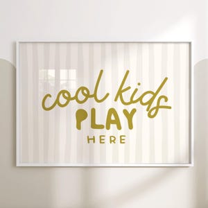 Może przedstawiać: Oprawiony wydruk z napisem „cool kids PLAY HERE” złotymi literami. Tło przedstawia pionowe paski w kolorze kremowym i jasnoszarym. Rama jest biała.