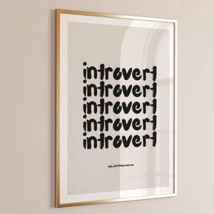 Puede incluir: Impresión artística enmarcada con la palabra "introvertido" repetida en negro. La frase "yep, all of them are me" está impresa en la parte inferior. El marco es dorado.