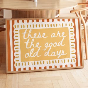 May include: A framed print featuring the phrase "these are the good old days" in white script on a yellow backdrop. The artwork has a decorative border and is set in a light wood frame. The print is leaning against a table.