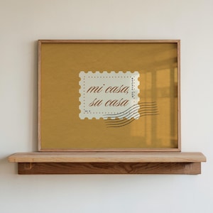 May include: A framed print with a mustard-yellow background and a postage stamp design. The stamp reads "mi casa, su casa" in a serif font. The frame is made of light-coloured wood, and the print is displayed on a wooden shelf.