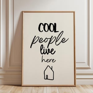 Könnte beinhalten: Gerahmter Druck mit den Worten "COOL people live here" in schwarzer Schrift. Darunter eine einfache Hausumrisszeichnung. Der Hintergrund ist cremefarben, der Rahmen hellbraun.