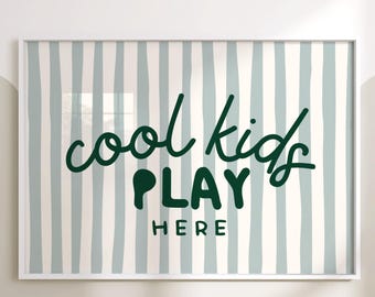 Op de afbeelding: Een ingelijste print met een witte rand toont de tekst "cool kids PLAY HERE" in donkergroen tegen een achtergrond van verticale strepen in lichtblauw en gebroken wit. Geschikt voor een kinderkamer of speelruimte.