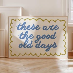 May include: A framed canvas print featuring the phrase "these are the good old days" in blue script. The text is on a cream background, framed by a scalloped green border, and set in a natural wood frame.