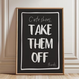 Op de afbeelding: Een ingelijste kunstdruk met een zwarte achtergrond en witte tekst. De tekst luidt "Cute shoes. TAKE THEM OFF thanks." De lijst is lichtbruin van kleur. De print wordt weergegeven op een houten vloer.