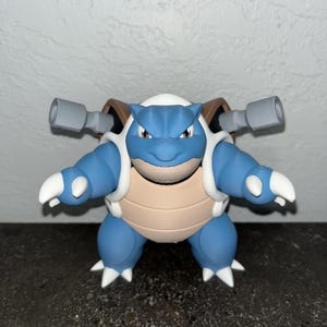 May include: A blue, tan, and white figurine of a Blastoise character. The figure has a blue body, white accents, and two gray cannons on its back. The figure is standing with its arms out.